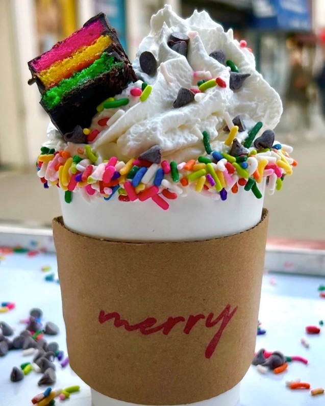 Merry Hot Cocoa cup topped with whipped cream, chocolate chips, a sprinkle-dipped rim and a rainbow cookie