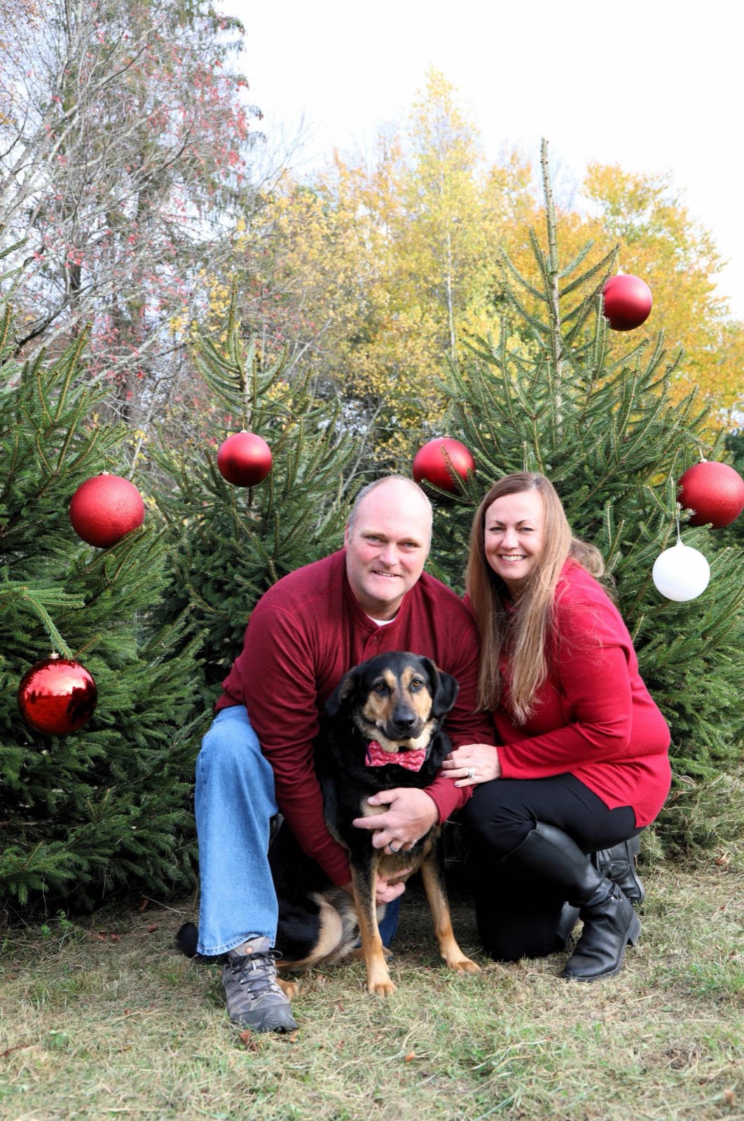 Couple in red with their dog in a bow tie among decorated trees and fall foliage