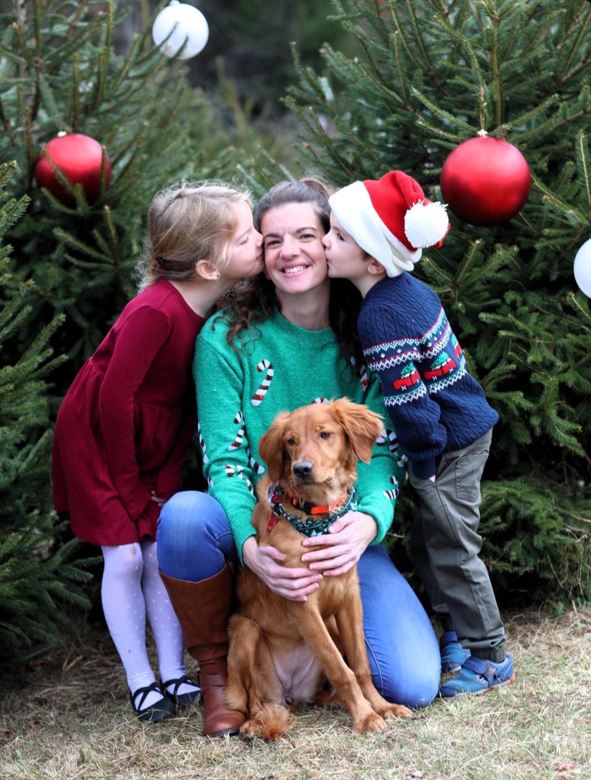 Two children kissing their mother while their golden retriever sits in front of decorated Christmas trees